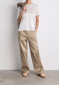 Biała koszulka z krótkim rękawem zestawiona z beżowymi spodniami cargo z bocznymi kieszeniami. Model stoi na betonowej podłodze, trzymając kurtkę.
