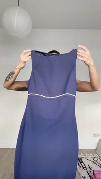 Personne avec des tatouages tenant une robe sans manches bleue marine ornée d'une rangée de perles autour de la taille dans une pièce minimaliste avec une lanterne en papier.