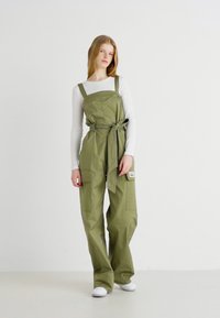 Oliven grøn overall jumpsuit med vide ben, justerbare stropper, en knyttet talje og sidelommer. Brukt over en tettsittende hvit langermet topp.