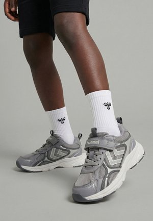 Ben iført sorte shorts, hvide sokker med bi-logo og grå Hummel-sneakers med velcrobånd på grå baggrund.