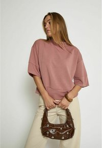 Top oversized rosa con maniche corte, abbinato a pantaloni beige. Borsa marrone con borchie argentate tenuta in mano. Tessuto di consistenza liscia.