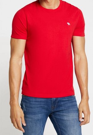 Man draagt een effen rood T-shirt met korte mouwen en een blauwe spijkerbroek, staand tegen een witte achtergrond.