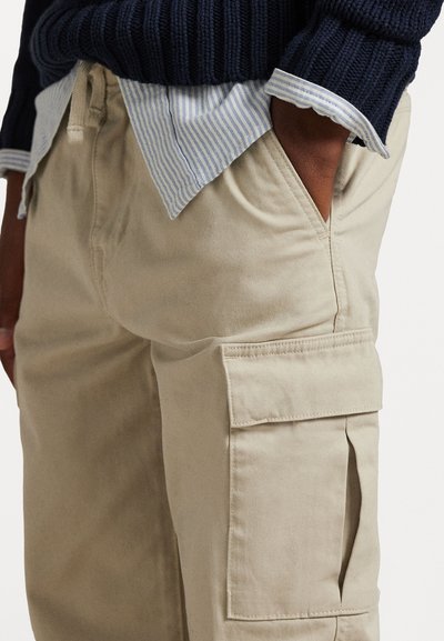 Μπεζ cargo παντελόνια από βαμβάκι, με πλευρικές τσέπες, χαλαρή εφαρμογή και λεία υφή, συνδυασμένα με πουκάμισο με ρίγες μπλε και λευκό.