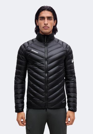 Mann mit dunklem Haar trägt eine schwarze Daunenjacke mit Frontreißverschluss und Logos auf Brust und Ärmel, steht vor hellem einfarbigem Hintergrund.