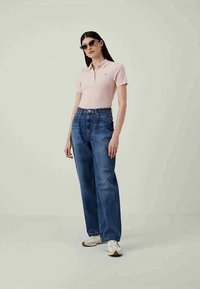 Jovem mulher a usar óculos de sol, uma camisola polo rosa clara, jeans azuis de cintura alta e ténis brancos, a posar contra um fundo simples.