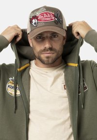 Hoodie beige avec des patchs de logo, t-shirt beige en dessous, et casquette en maille beige avec des designs brodés colorés et une sangle réglable.