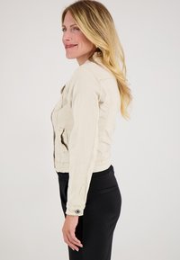 Blonde vrouw in een beige jas en zwarte broek staat in profiel, glimlachend tegen een effen witte achtergrond.