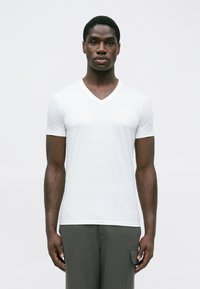 Άνδρας που φοράει ένα απλό λευκό T-shirt με V και σκούρους γκρι παντελόνες, στέκεται μπροστά από έναν απλό λευκό τοίχο, κοιτώντας μπροστά.