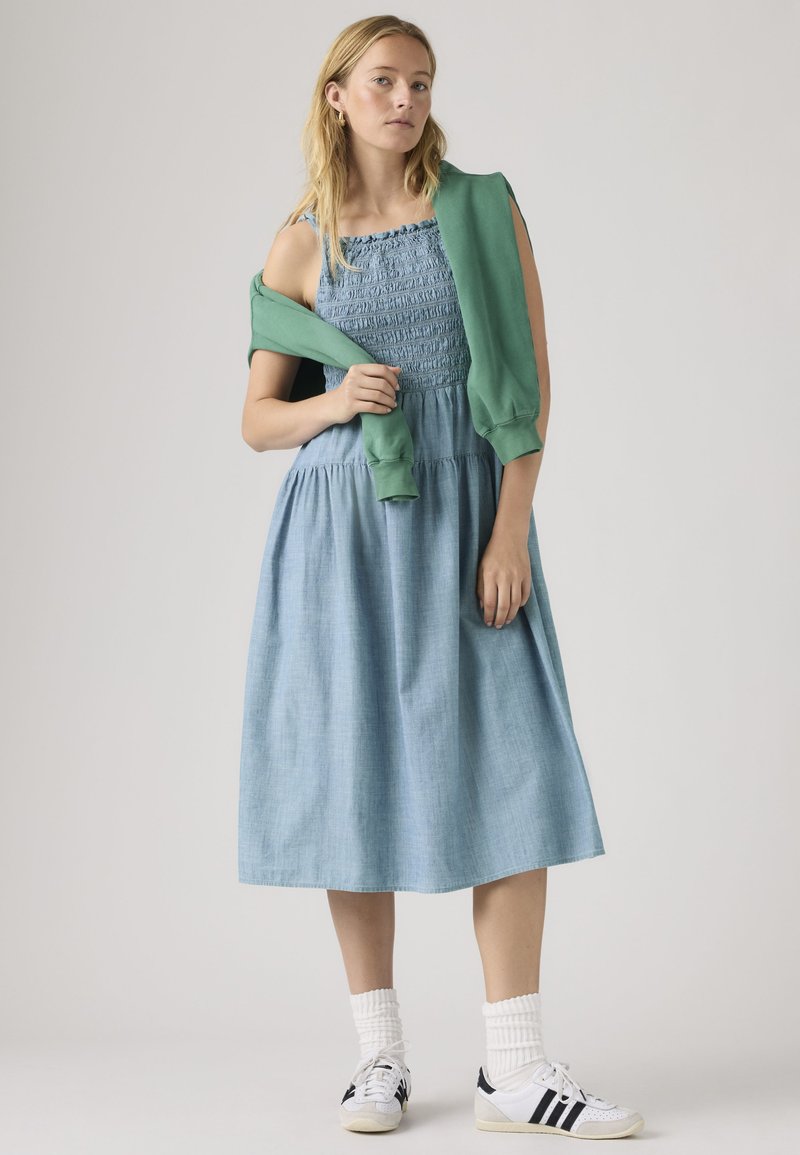 Femme aux longs cheveux blonds portant une robe sans manches bleu clair, des chaussettes blanches, des baskets blanches et un pull vert posé sur les épaules.