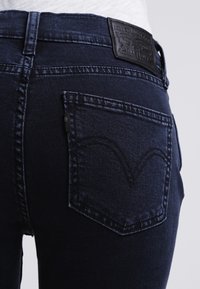 Acercamiento de unos pantalones vaqueros oscuros que muestran el bolsillo trasero con un diseño cosido y una etiqueta de cuero en la cinturilla.