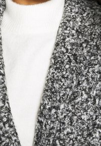 Grå och vit texturerad cardigan med ett mjukt, krusigt stickat mönster, lager på lager över en vit ribbad polokrage.