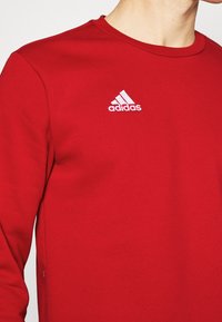 Rode Adidas sweatshirt met een ronde halslijn, gemaakt van een gestructureerde stof, met een wit logo op de linkerborst. Eenvoudig ontwerp, geen patronen.