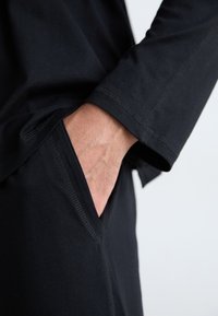 Chemise noire à manches longues avec une texture lisse et des coutures visibles. Une main repose dans la poche avant d'un pantalon noir assorti.