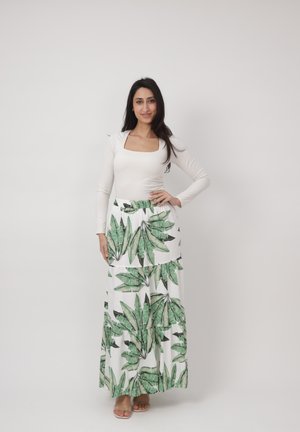 Femme debout, main sur la hanche, portant un haut blanc à manches longues et une jupe longue blanche à imprimé feuilles vertes, fond neutre.