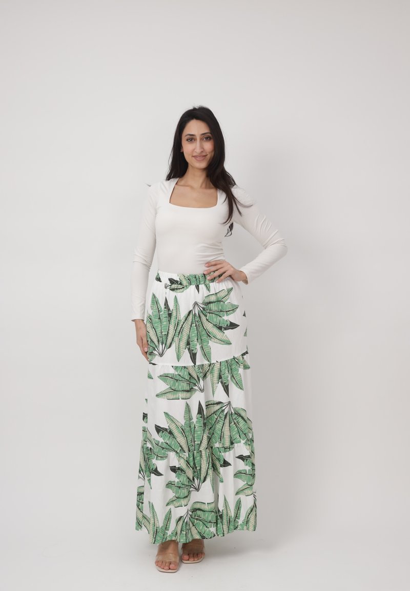 Femme debout, main sur la hanche, portant un haut blanc à manches longues et une jupe longue blanche à imprimé feuilles vertes, fond neutre.