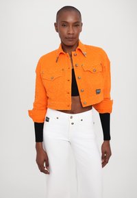 Giacca di jeans cropped arancione brillante con superficie testurizzata, chiusura con bottoni metallici, due tasche sul petto e cuciture a contrasto. Abbinata a pantaloni bianchi.