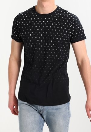 Homme portant un t-shirt noir à manches courtes avec de petits motifs géométriques blancs, associé à un jean bleu clair, debout devant un fond uni.
