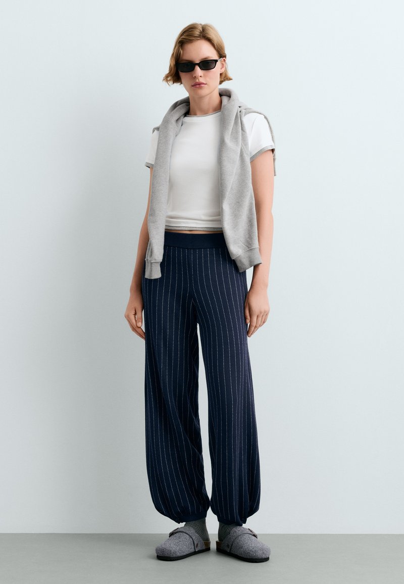 Femme portant des lunettes de soleil noires, un t-shirt court blanc, un sweat gris posé sur les épaules, un pantalon bleu marine à fines rayures, des chaussettes grises et des chaussons gris.