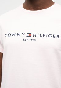 Ροζ βαμβακερό t-shirt με στρογγυλή λαιμόκοψη που φέρει το λογότυπο "TOMMY HILFIGER" σε σκούρο μπλε χρώμα, με έμφαση σε ένα κόκκινο και μπλε ορθογώνιο. Κάτω από το λογότυπο αναγράφεται "EST. 1985".