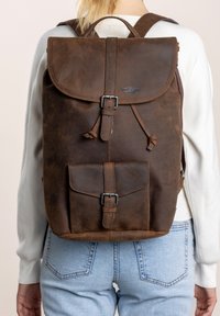 Mochila de cuero marrón con tapa plegable, cierre de hebilla de metal, bolsillo frontal y correas ajustables. Textura suave, con mínima visibilidad de la marca.