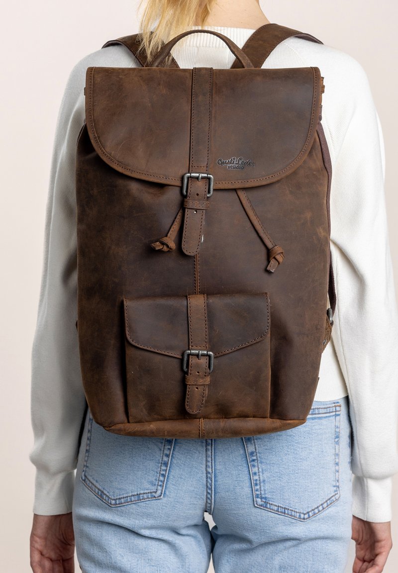 Mochila de cuero marrón con tapa plegable, cierre de hebilla de metal, bolsillo frontal y correas ajustables. Textura suave, con mínima visibilidad de la marca.