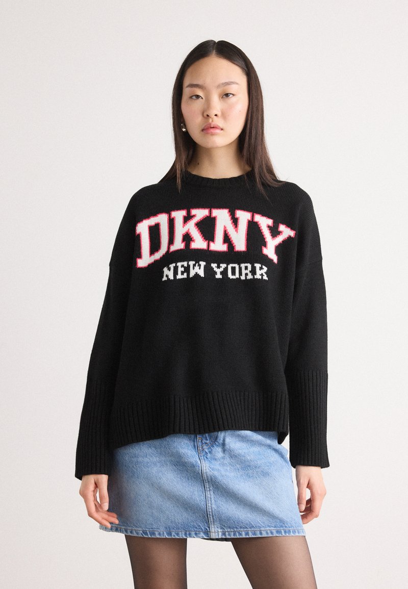Černý pletený svetr s žebrovaným lemem a manžetami, s nápisem "DKNY NEW YORK" ve velkých červených a bílých písmenech přes hrudník. V páru s denimovou sukní.