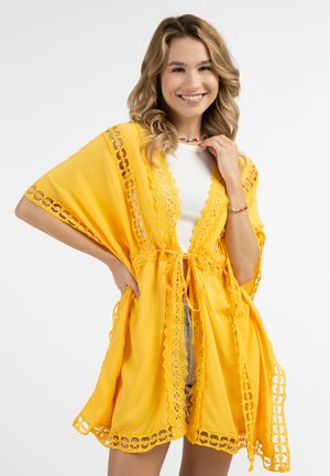 Copertura in stile kimono gialla con rifiniture in pizzo e apertura frontale, dotata di chiusura con lacci. Il tessuto leggero scorre liberamente ai bordi.