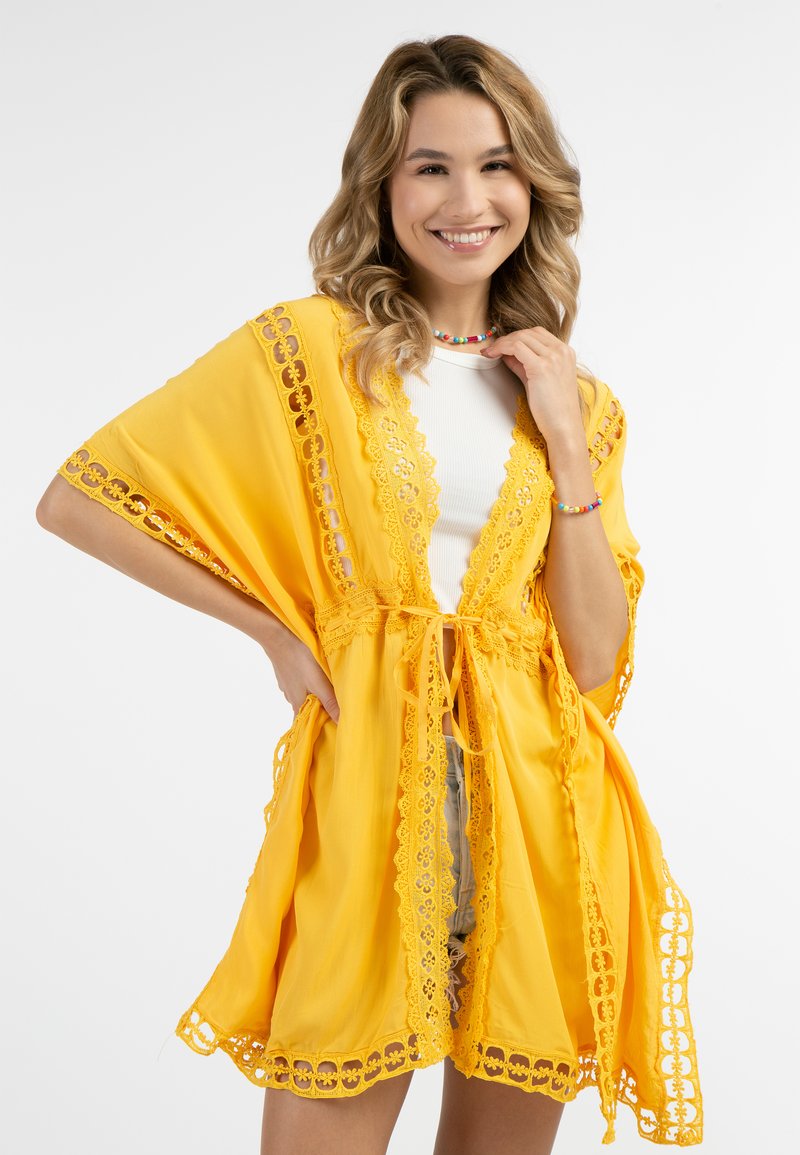 Keltainen kimono-tyylinen peite, jossa on pitsireunus ja avoin etuosa, sekä solmittava kiinnitys. Kevyt kangas laskeutuu väljästi reunoilta.