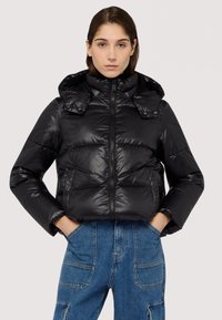 Giacca a vento nera con finitura lucida, colletto alto e chiusura con zip. Presenta tasche laterali e un cappuccio, indossata sopra pantaloni in denim blu.