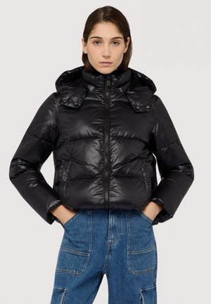 Sorte puffjakke med en blank finish, høj krave og lynlåslukning. Har sidelommer og hætte, båret over blå denimbukser.