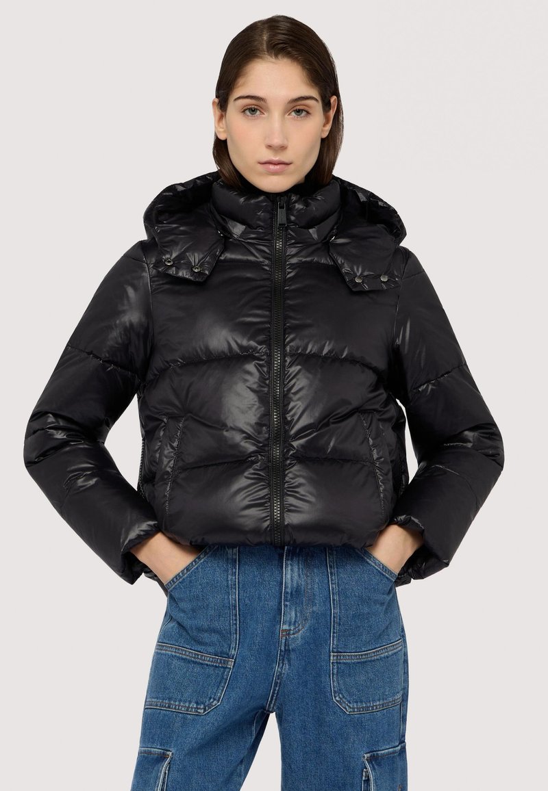 Giacca a vento nera con finitura lucida, colletto alto e chiusura con zip. Presenta tasche laterali e un cappuccio, indossata sopra pantaloni in denim blu.