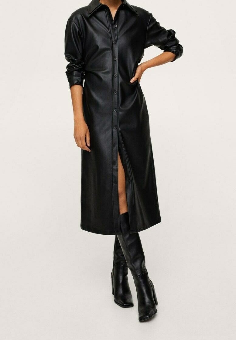 Femme portant une longue robe-chemise noire en similicuir boutonnée, manches retroussées, associée à des bottes noires à talons hauts montant aux genoux.