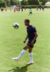 En manlig fotbollsspelare i en marinblå Puma-tröja och svarta shorts utför en bollkontrolleringsövning på gräs. Vita fotbollssockor och rosa skor bärs.