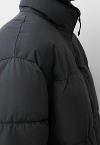 Gros plan d'une personne portant une veste d'hiver matelassée noire avec un col montant et des coutures visibles.