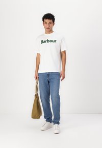 Bílý tričko s krátkým rukávem s logem "Barbour" v zelené barvě, kombinované s modrými džíny a bílými teniskami, držící béžovou kabelku přes rameno.