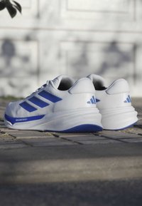 White athletic shoes with blue accents, featuring a textured upper, three-stripe design, cushioned sole, and pronounced heel.
