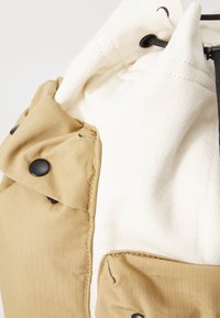 Beige og hvit ryggsekk laget av teksturert stoff, med snøring, trykknappdetaljer og en strukturert design.