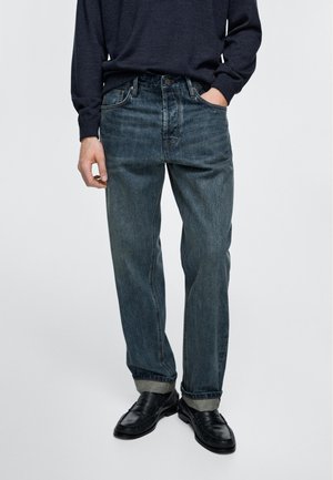 Homme portant un pull bleu foncé, un jean délavé bleu clair ample retroussé aux chevilles, et des mocassins en cuir noir, debout avec une main dans la poche.