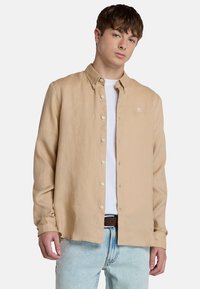 Camicia button-up beige in tessuto leggero, con maniche lunghe e un logo sul petto. Indossata sopra una t-shirt bianca, abbinata a jeans azzurri chiari.