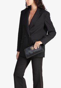Blazer strutturato nero con rever a punta, abbinato a pantaloni neri. Tiene una pochette nera texturizzata con chiusura in tonalità oro.