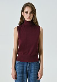 Burgunderfarbener ärmelloser Rollkragenpullover aus weichem Stoff, mit tailliertem Schnitt und glatter Textur, kombiniert mit blauen Jeans im weit geschnittenen Stil.