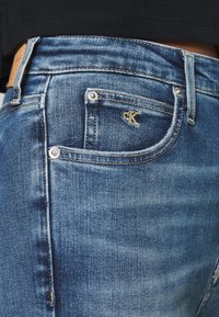 Jeans z denimu w średnim niebieskim odcieniu z subtelnym wyblaknięciem. Posiadają logo CK w pobliżu tylnej kieszeni, srebrne nity i standardowy pięciokieszeniowy design.