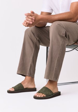 Muškarac sjedi na stolici, nosi bež hlače i zelene sandale otvorenih prstiju s remenom preko stopala.