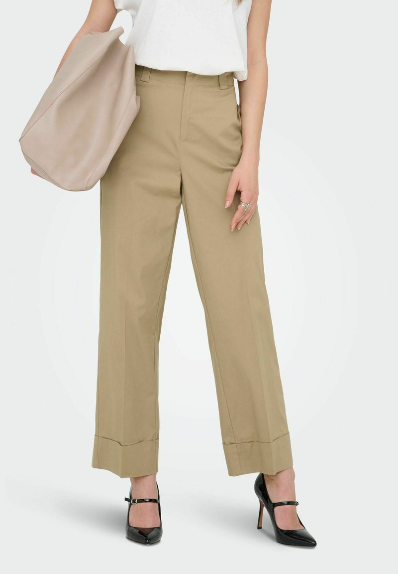 Femme portant un pantalon large beige taille haute, un haut sans manches blanc, des escarpins Mary Jane noirs, tenant un grand sac fourre-tout beige à un bras.
