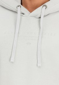 Gaišpelēka kapuce, kas izgatavota no mīkstas auduma ar teksturētu virsmu. Iekļauj baltus aukliņas un smalku zīmolu apdruku priekšpusē.