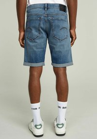 Shorts en denim bleu clair avec un ourlet retroussé, dotés de deux poches arrière et d'une étiquette de marque noire. Portés avec des chaussettes blanches et des chaussures blanches.