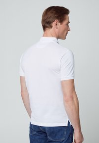 White polo shirt with short sleeves, collared neck, side slits, and smooth cotton texture, worn with blue denim jeans.
