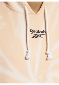 Pale peach hoodie with white drawstrings, featuring a tie-dye pattern. Reebok logo embroidered in black on the chest area. Soft fabric texture.