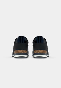 Une paire de baskets noires avec des accents en liège au talon et des semelles blanches, vues de l'arrière sur un fond blanc uni.