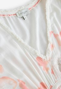 Hvid blomsterkjole med bløde lyserøde mønstre, med V-udskæring og frynsekant. Etiketten lyder "By Ted Baker, London," størrelse S.